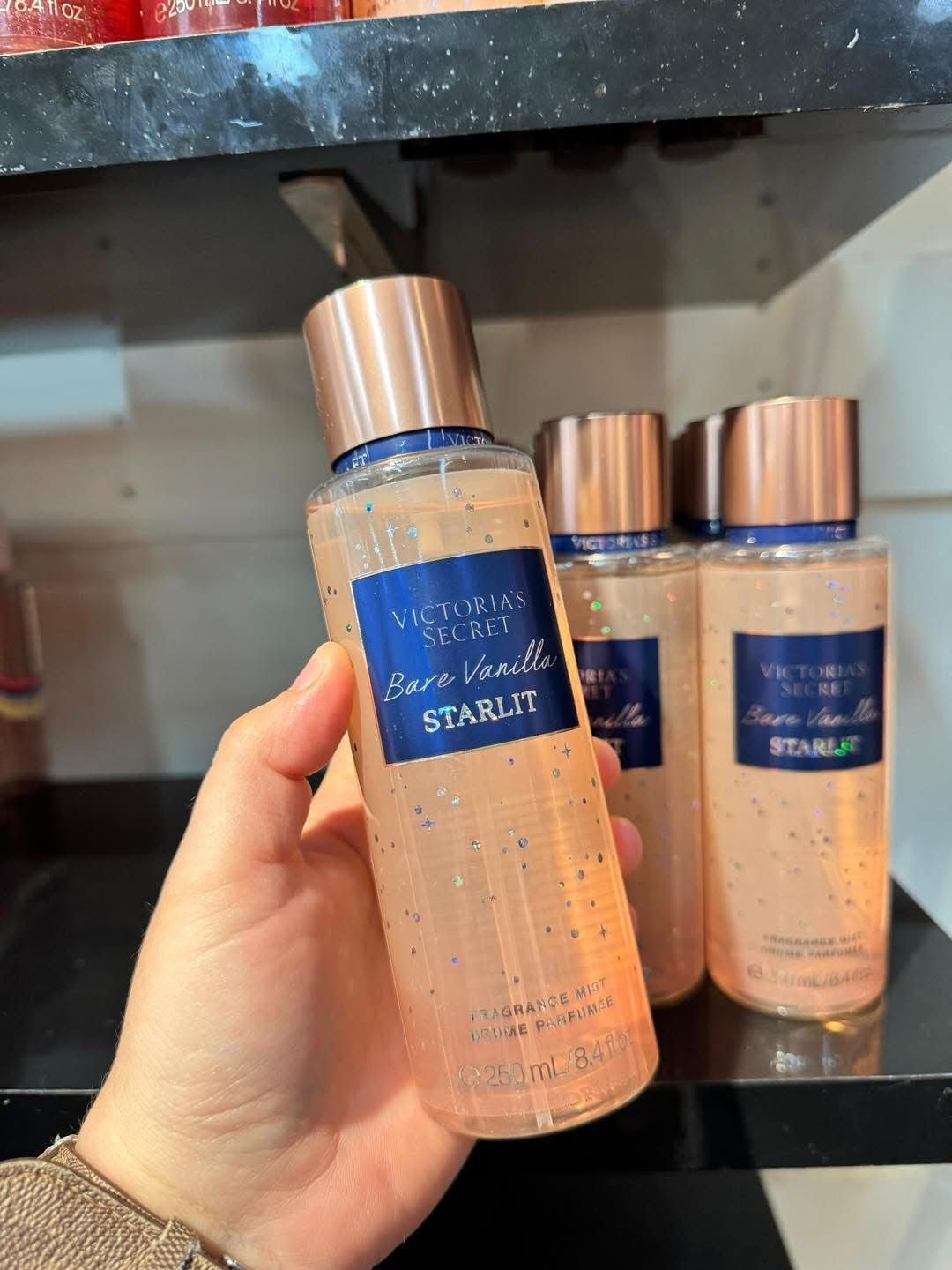 Uma mão segurando um frasco transparente de victoria’s secret bare vanilla starlit com tampa rosé e líquido pêssego glitterado