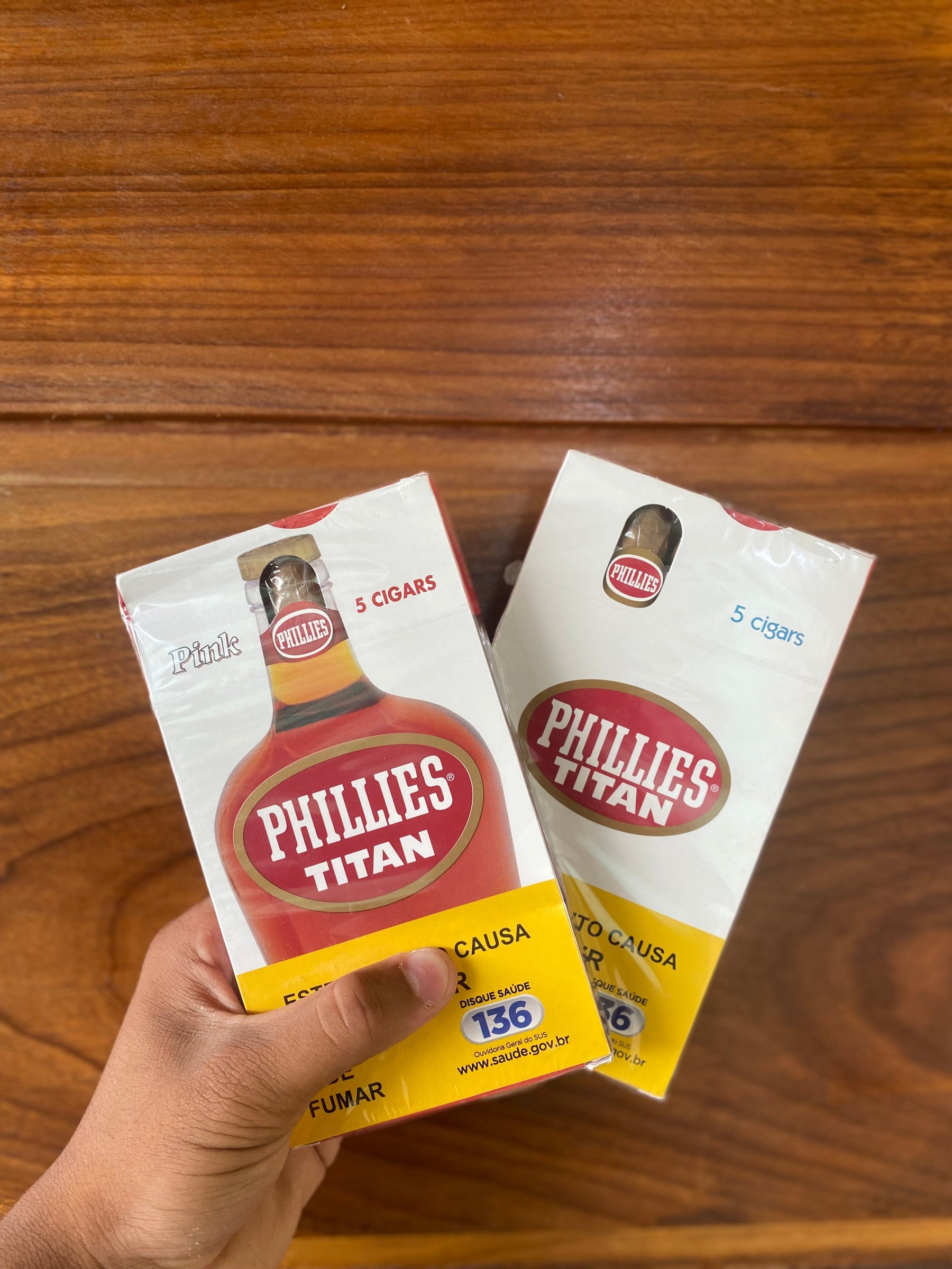 Duas embalagens de charutos phillies titan tradicional em uma mão, uma rosa e outra comum, sobre superfície de madeira