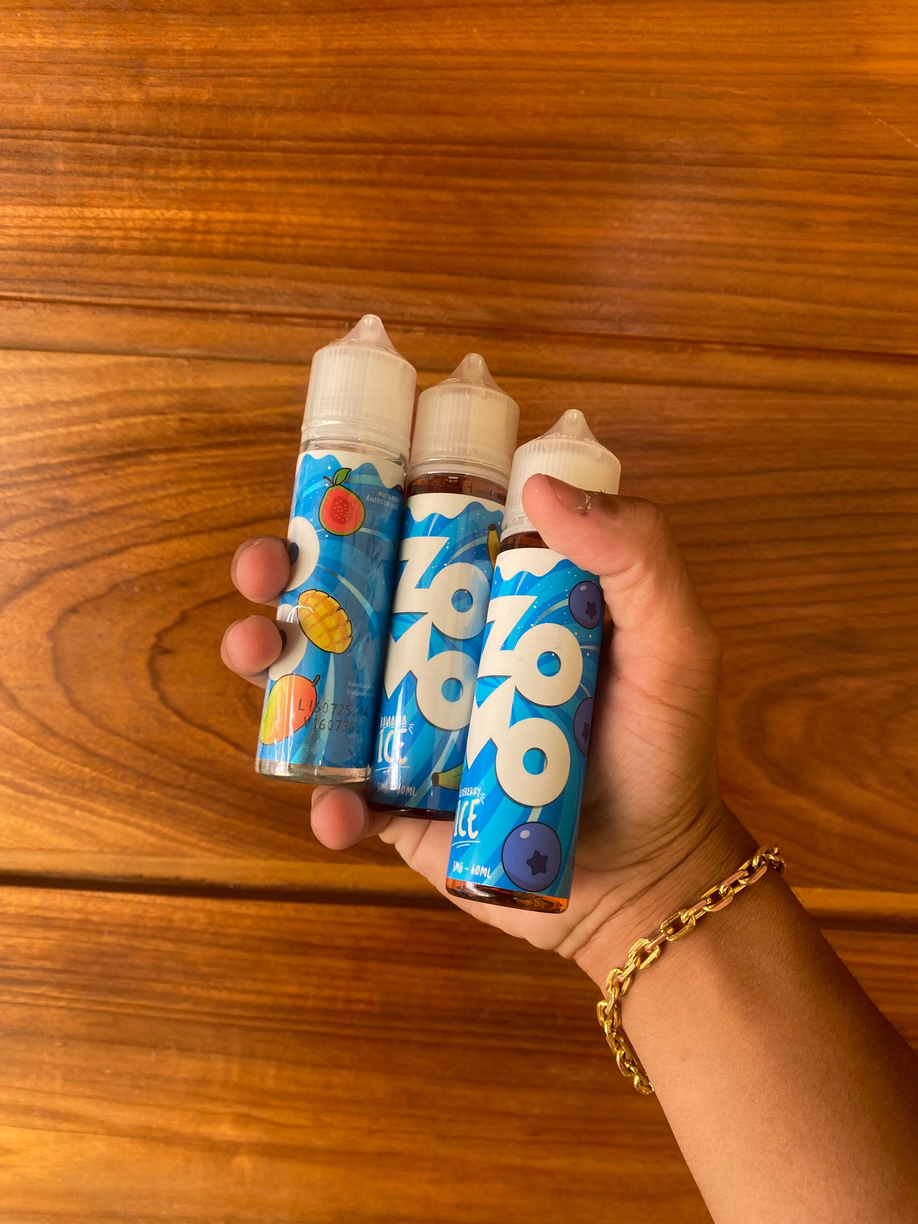 Três frascos de e-líquido juice zomo 60ml 3mg azul e branco com ilustrações de frutas em uma mão contra superfície de madeira