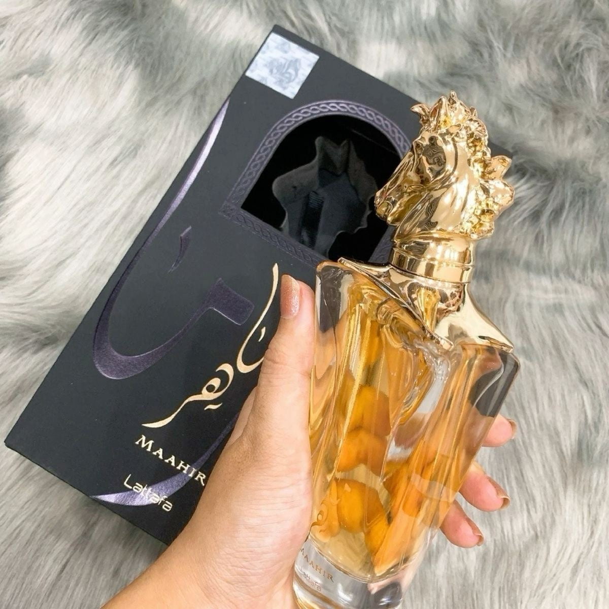 Perfume maahir lattafa gold 100ml em frasco de vidro facetado âmbar com tampa dourada em forma de cabeça de cavalo ao lado da caixa preta